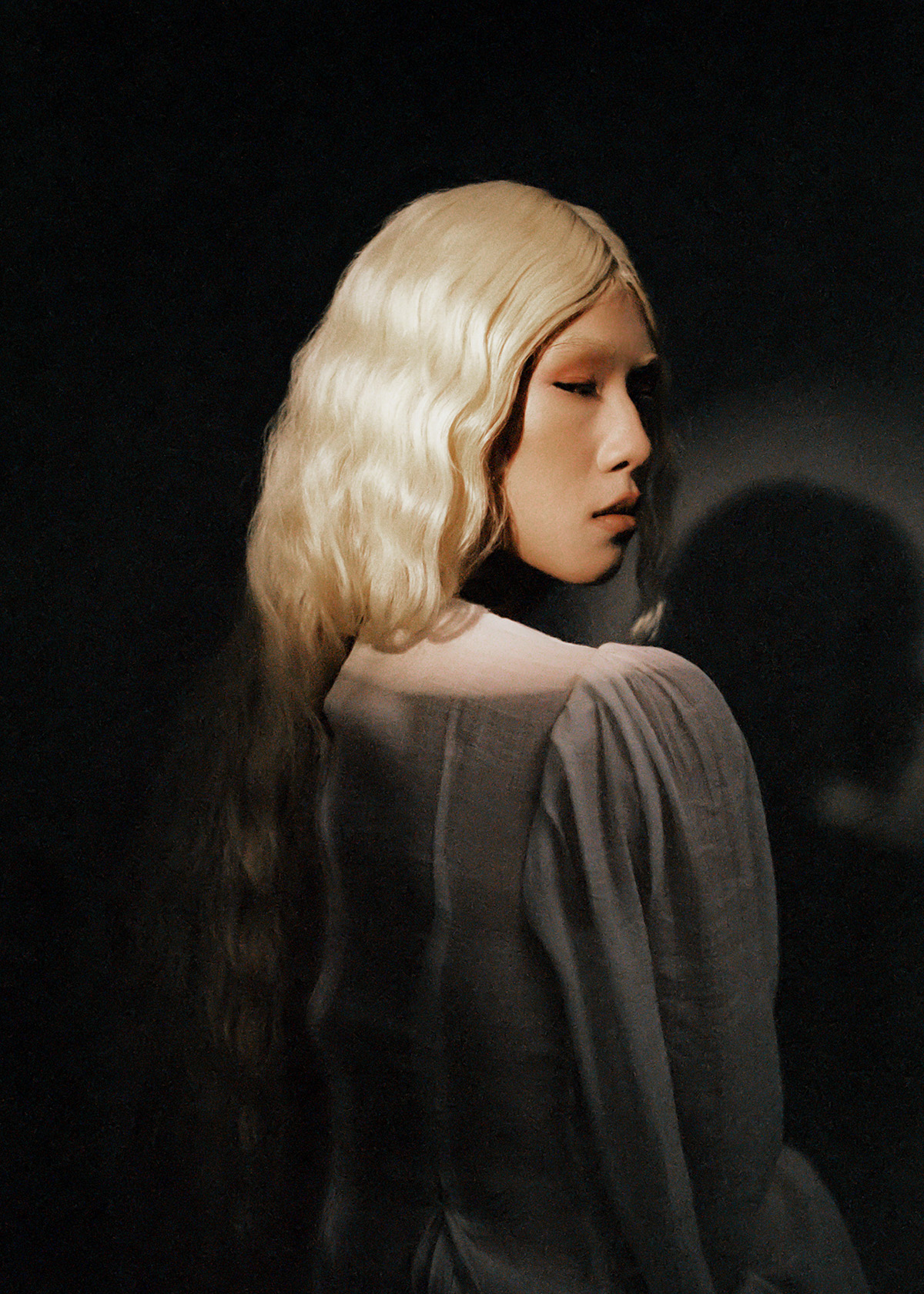 Portrait of an Asian woman with blonde wavy hair looking to the right, wearing a sheer grey top against a dark background.
