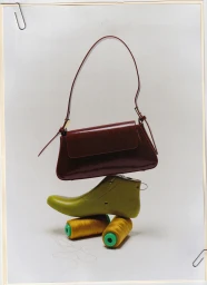 A still life studio photograph by Jess Brohier featuring a stacked composition with a brown leather handbag, a green shoe last, and two yellow spools.