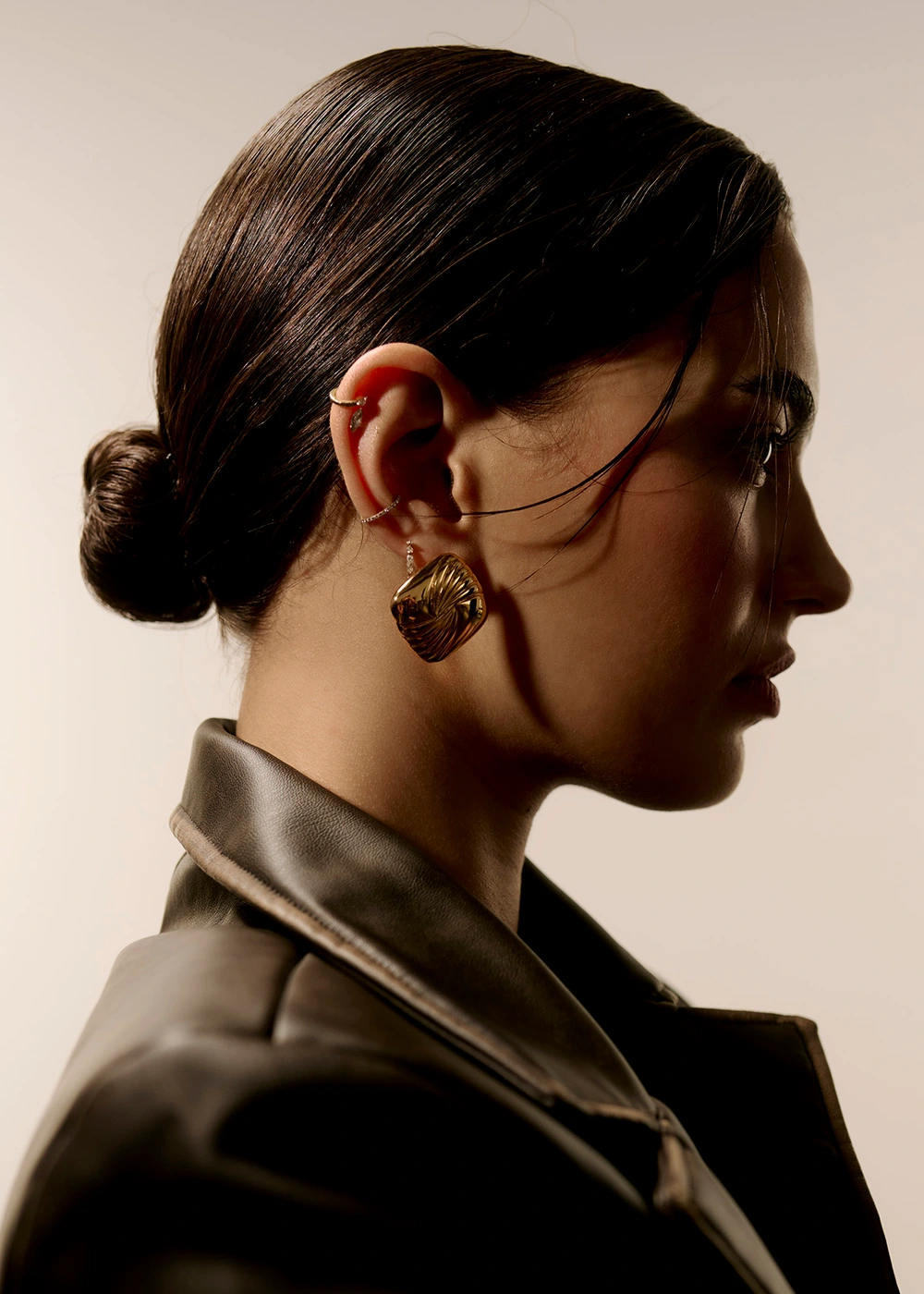 Profile portrait of a woman wearing gold earrings and a dark leather jacket against a light background by photographer Jess Brohier.