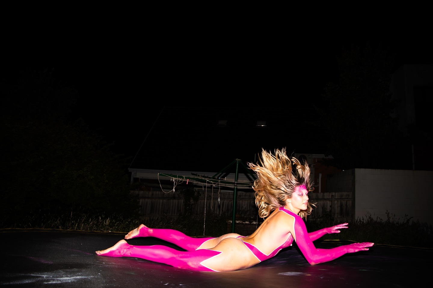 Woman with pink body paint in dynamic pose on trampoline, illuminated by a single light source at night, creating a striking contrast.