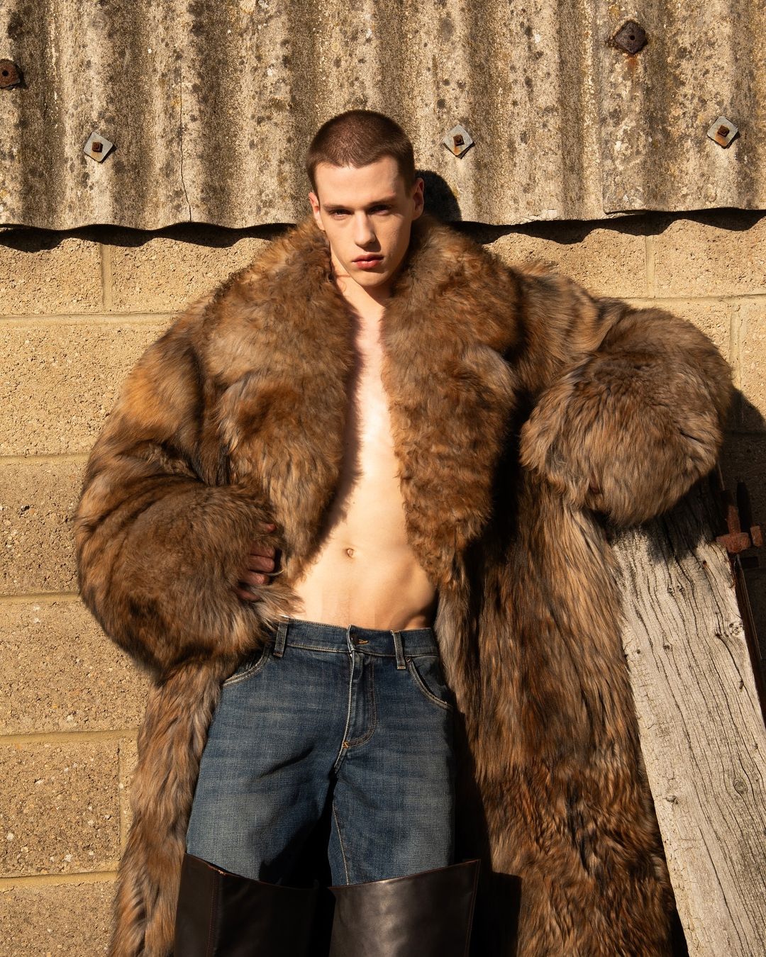 Man in a large fur coat poses against an exterior wall, showing off his abs and jeans. Leather boots are also visible.