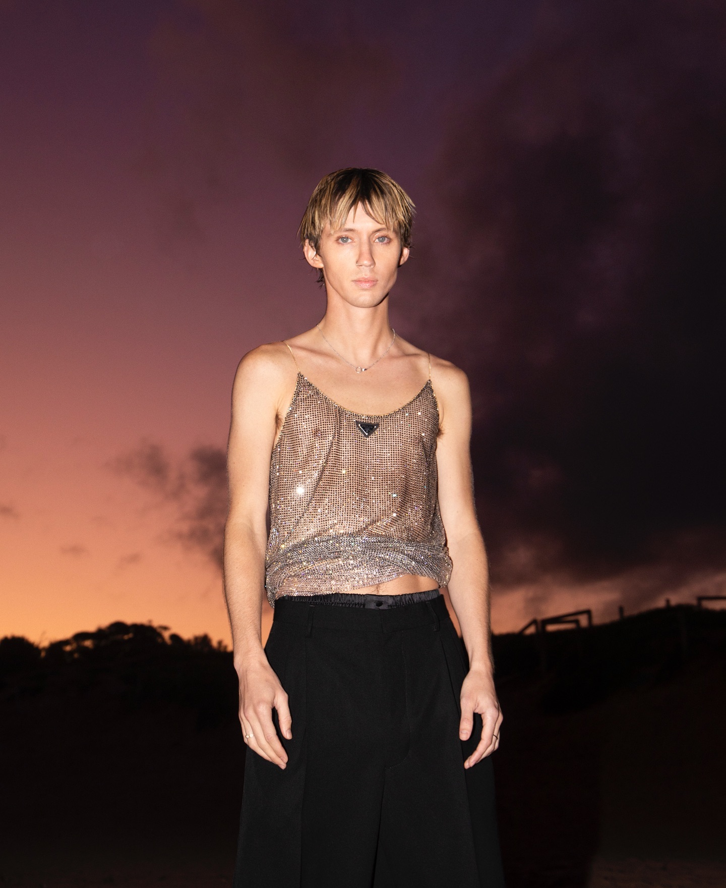 Young adult poses outdoors at sunset in silver sequin tank top and black pants against a dramatic, colorful sky.