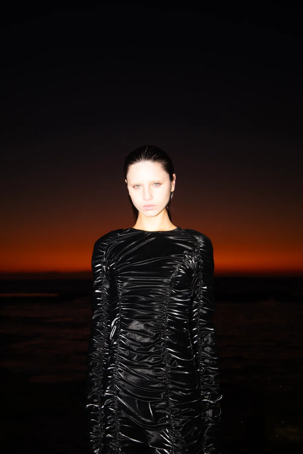 A young woman in a black ruched dress poses outdoors with an orange sunset background, creating a dark yet striking fashion portrait.