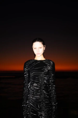 A young woman in a black ruched dress poses outdoors with an orange sunset background, creating a dark yet striking fashion portrait.