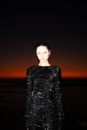 Model in a black ruched dress standing against a dark sunset horizon over water, photographed by Zac Bayly.