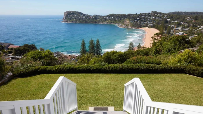 High-angle view from a grassy terrace down white stairs toward a sandy beach, blue ocean, and coastal headland.