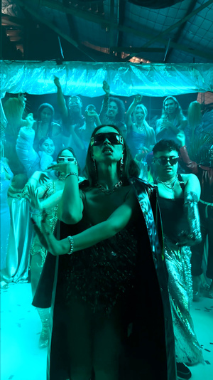 A woman in sunglasses and a long coat poses in front of a crowd with blue lighting, creating a vibrant and energetic atmosphere.