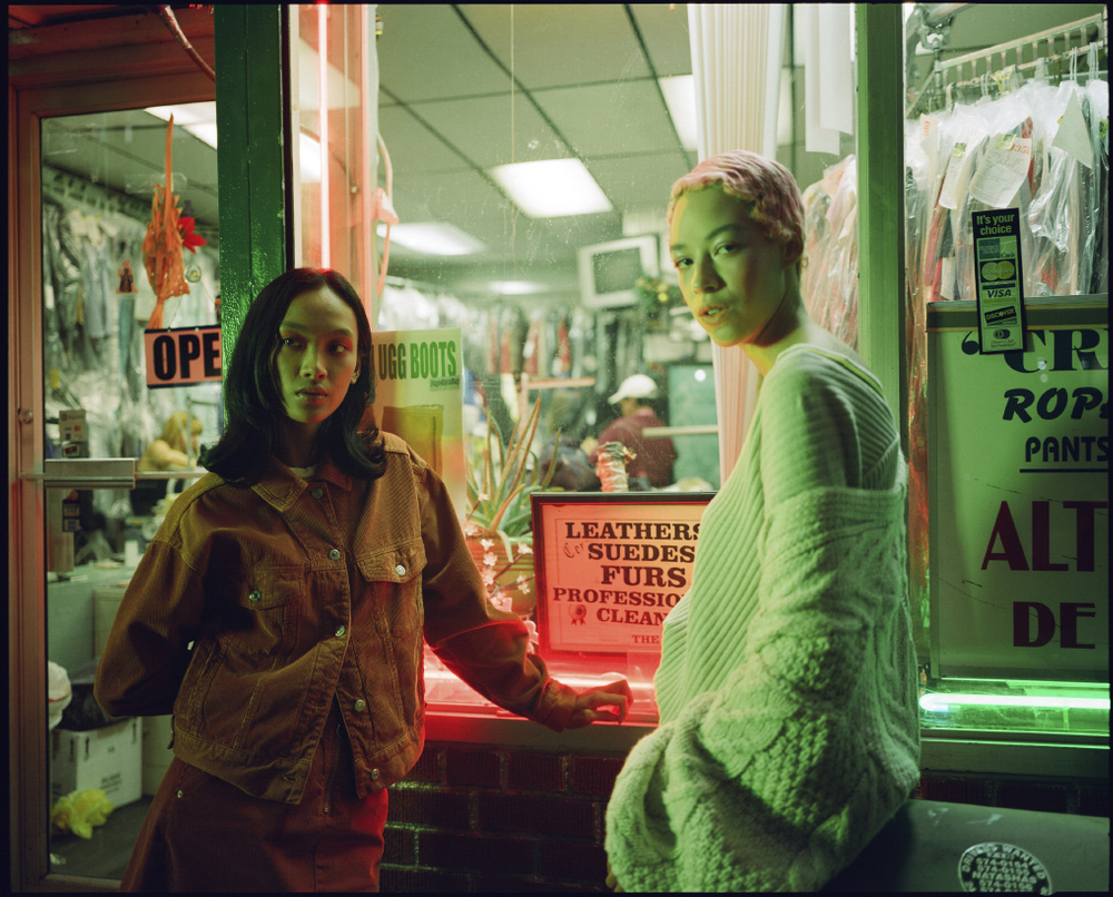 Two women pose near a storefront at night, illuminated by neon signs and warm indoor lighting, wearing fashionable clothing.