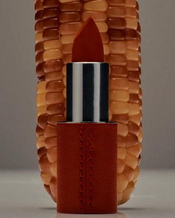 A lipstick is placed in front of a corn cob, creating a beauty advertisement, with a unique and creative presentation in a studio setting.