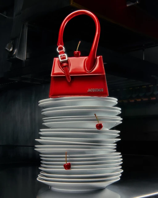 Red patent leather Jacquemus handbag on a stack of white plates, adorned with cherries. Fashion and accessories concept.