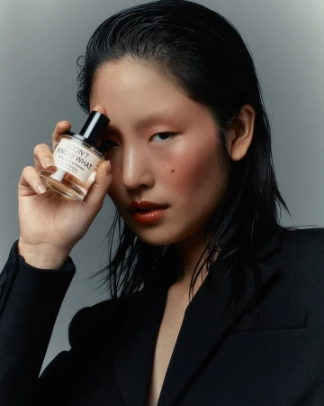 East Asian woman holding a perfume bottle near her face wearing a black blazer against a grey backdrop