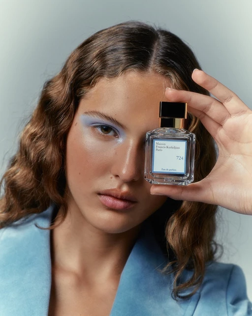 Woman with curly brown hair is holding up a glass perfume bottle with a black cap in a close-up studio shot.