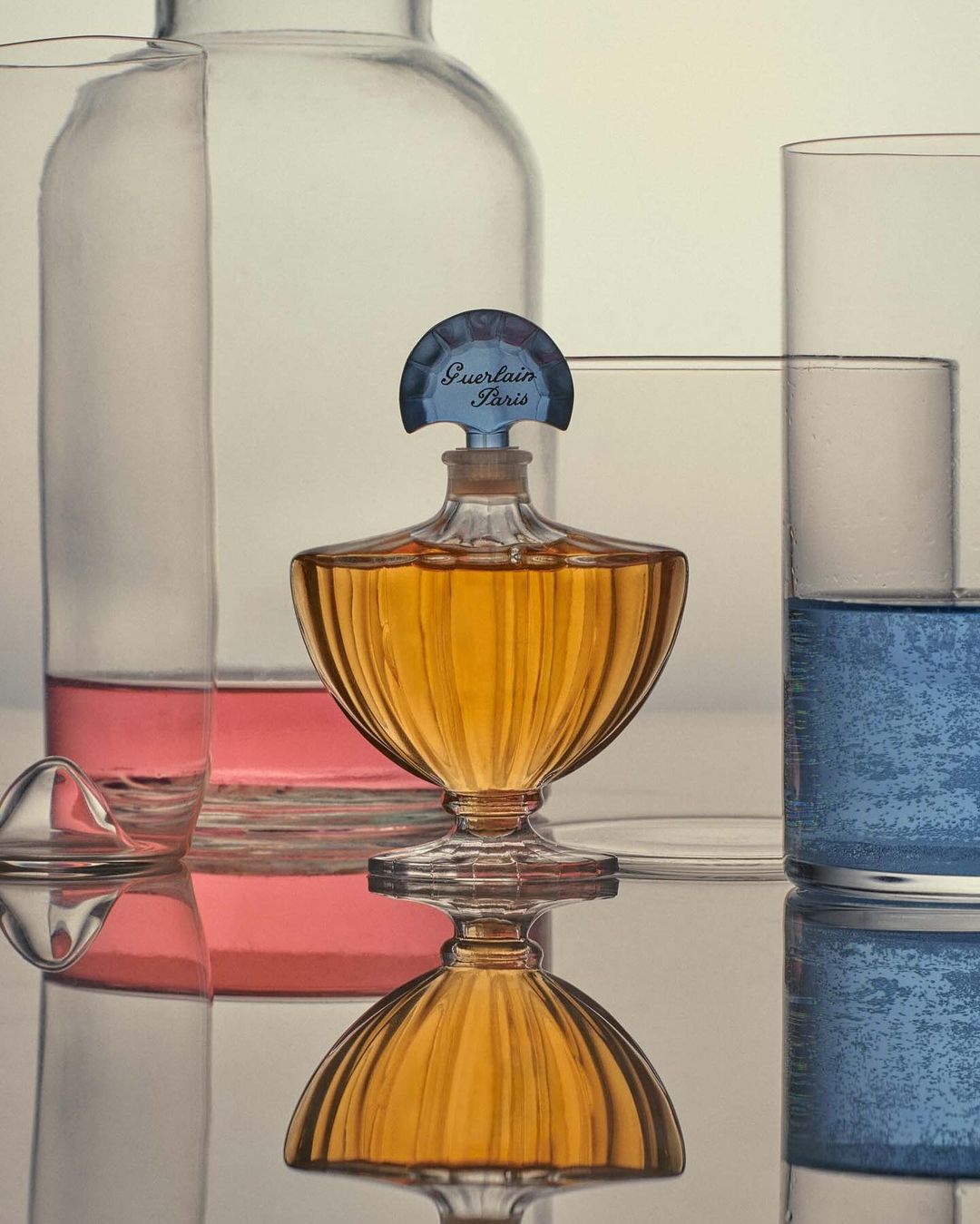 Still life close-up of vintage Guerlain perfume bottle on a reflective surface with clear laboratory beakers in the background.