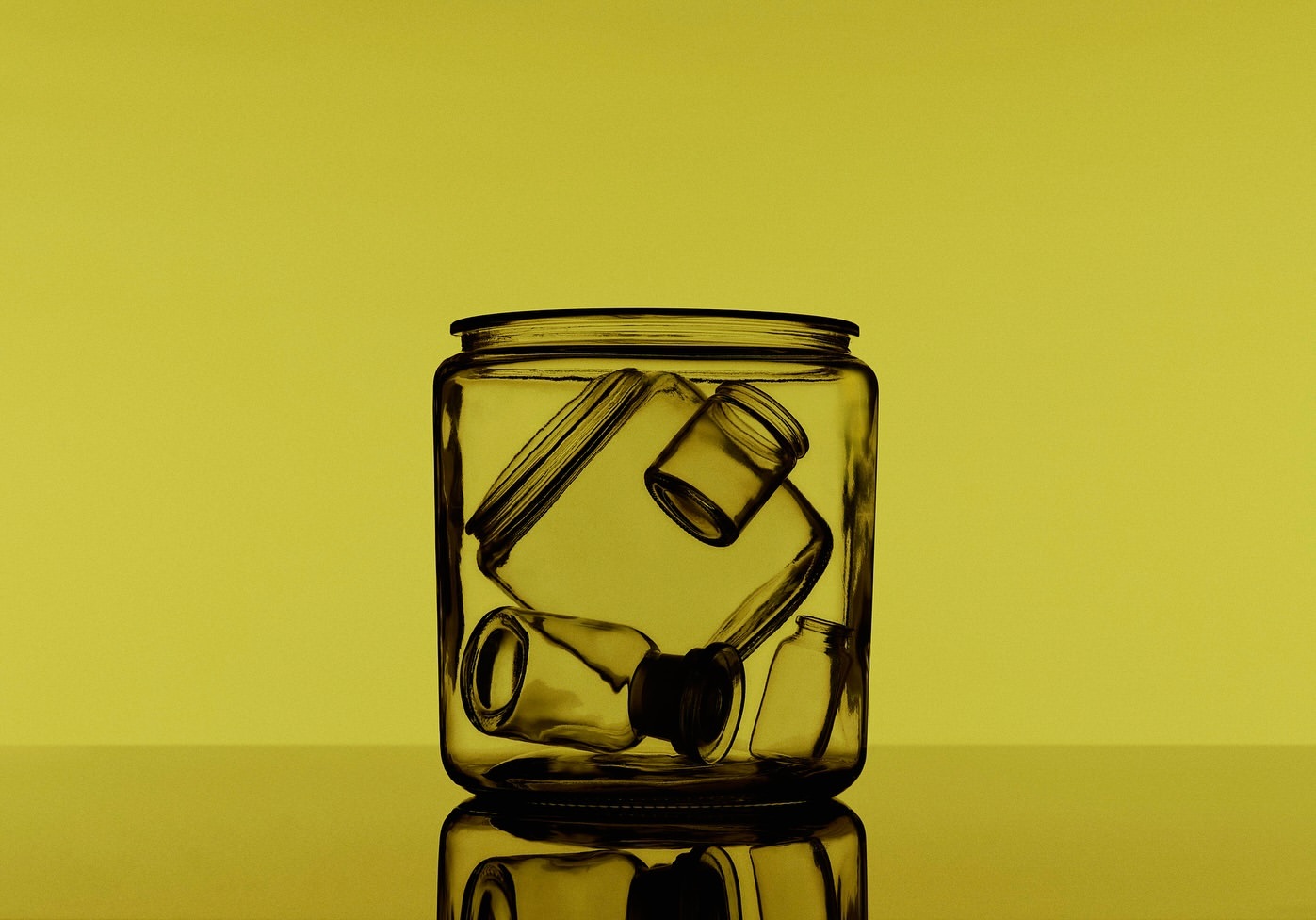 Still life shot of a clear glass jar containing an assortment of small glass bottles and containers against a solid yellow background.