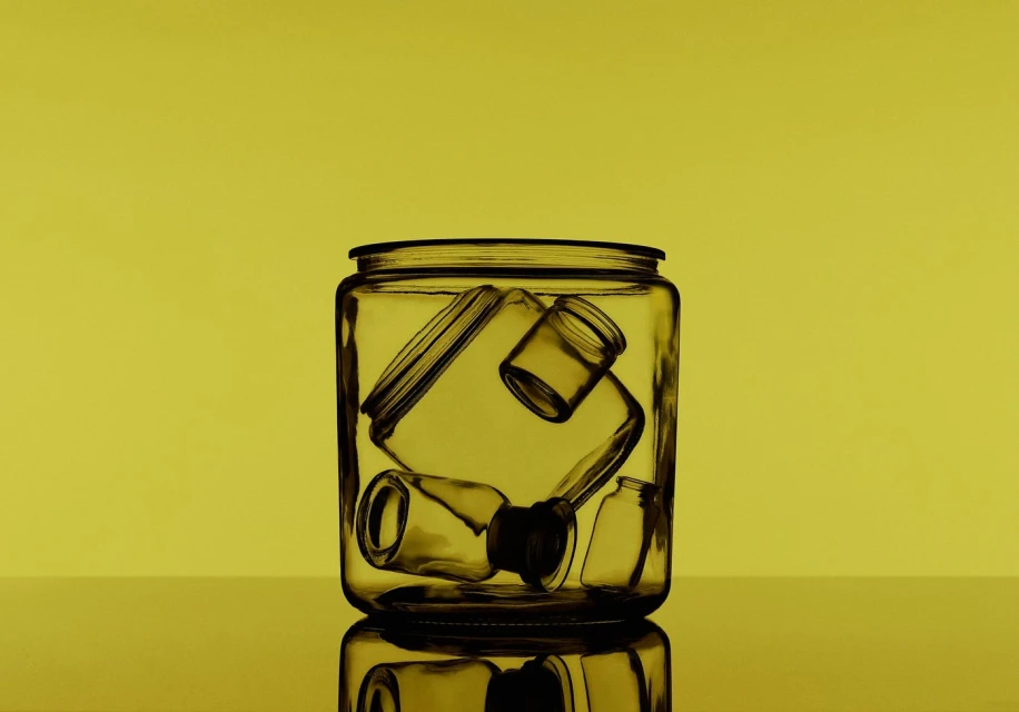 Still life shot of a clear glass jar containing an assortment of small glass bottles and containers against a solid yellow background.