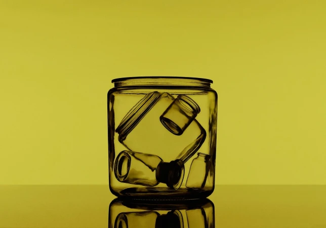 Still life shot of a clear glass jar containing an assortment of small glass bottles and containers against a solid yellow background.