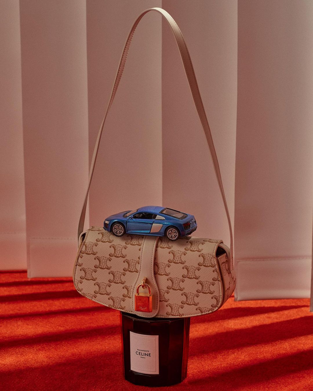 Designer handbag with logo pattern and a blue toy car perched on top, placed on a candle against a peach and red background.