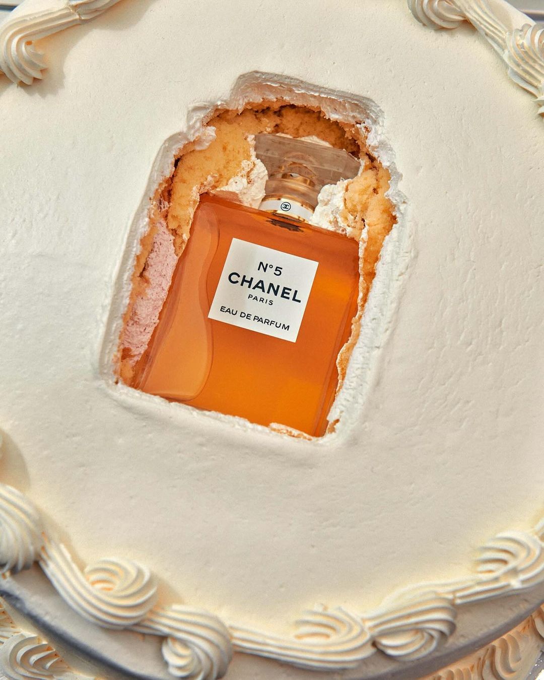 Elevated view of a square white cake with decorative frosting, featuring a Chanel No. 5 perfume bottle inside a carved-out section.