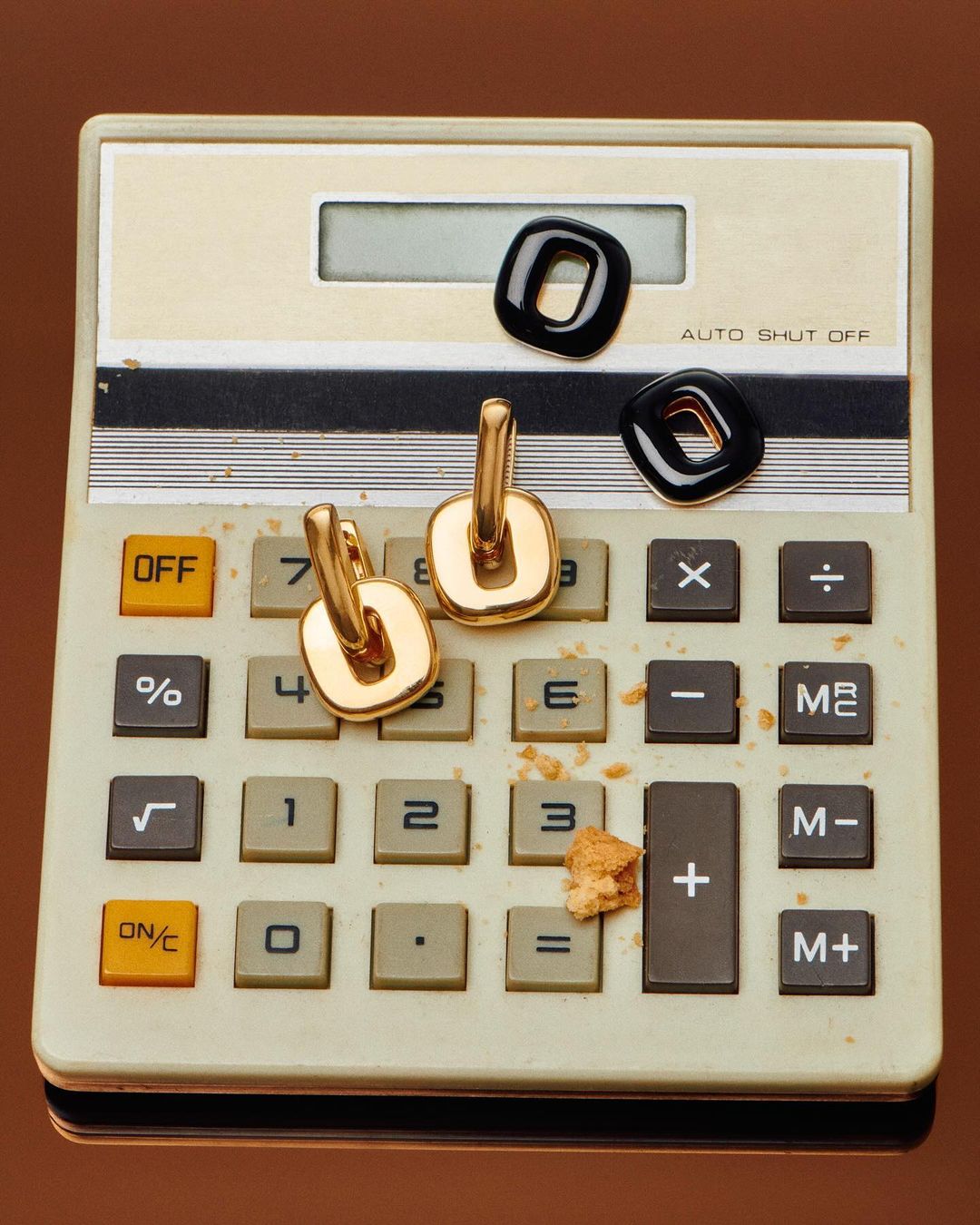 Gold earrings and black stone rings styled on a beige calculator with cookie crumbs, set against a brown background.