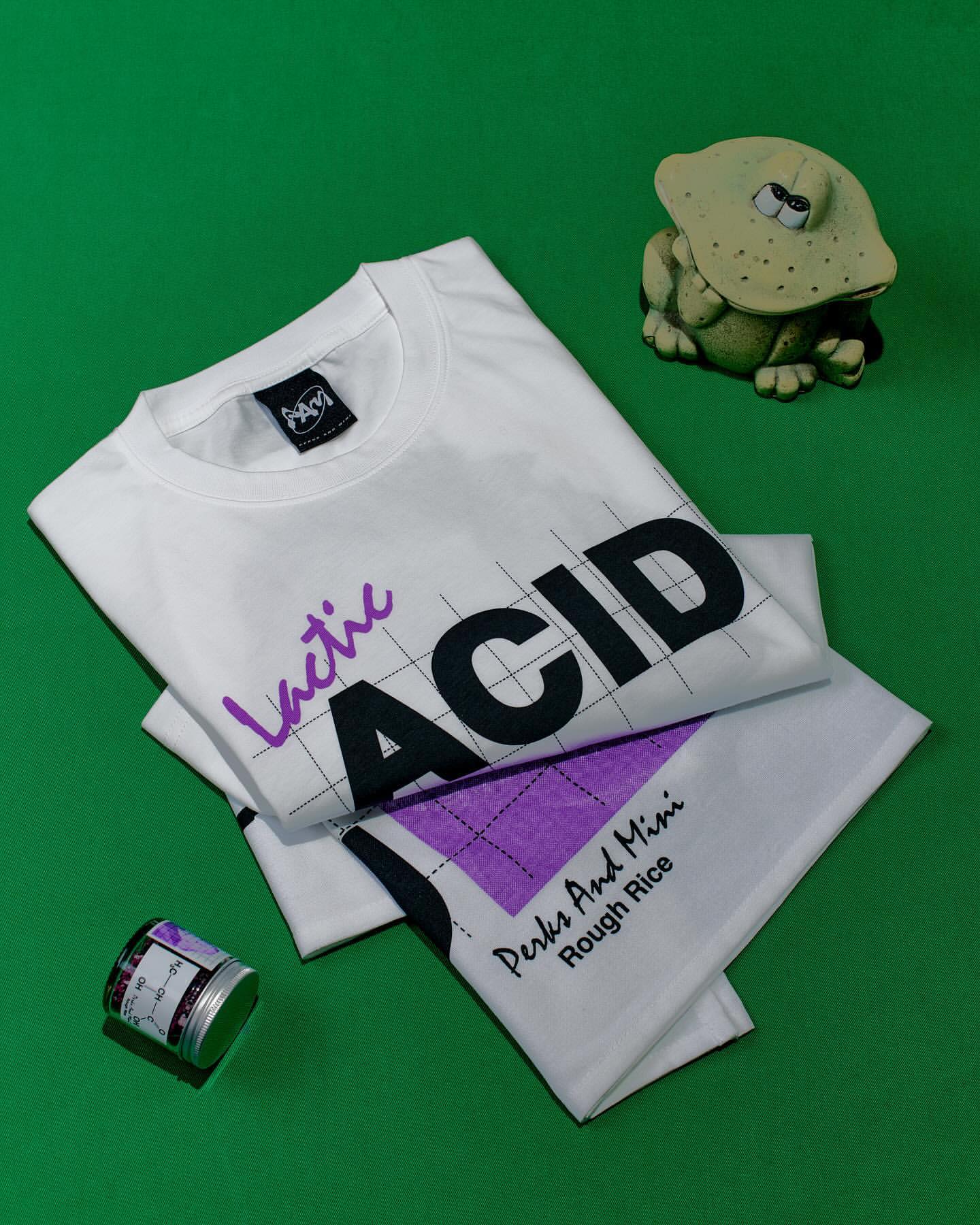 Still life photo of a folded white t-shirt with text designs on a green background, next to a frog figurine and a small container.