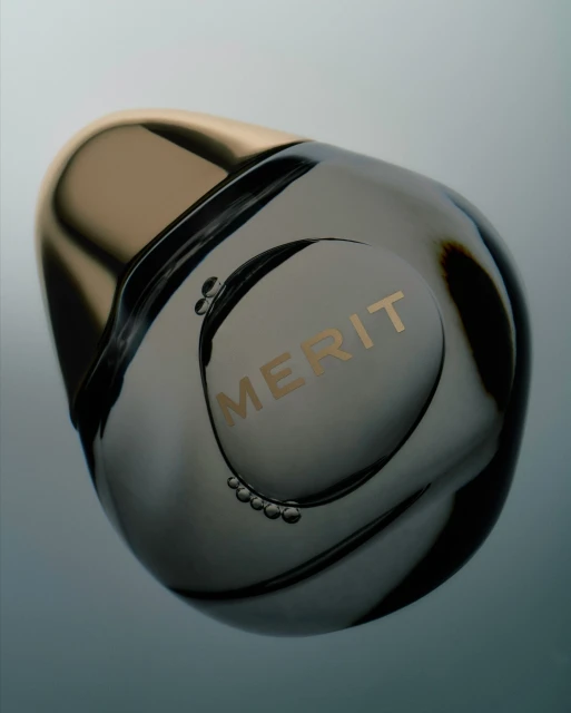 Elevated product shot of a Merit beauty product in a round, transparent container with a gold lid on a soft gradient background.