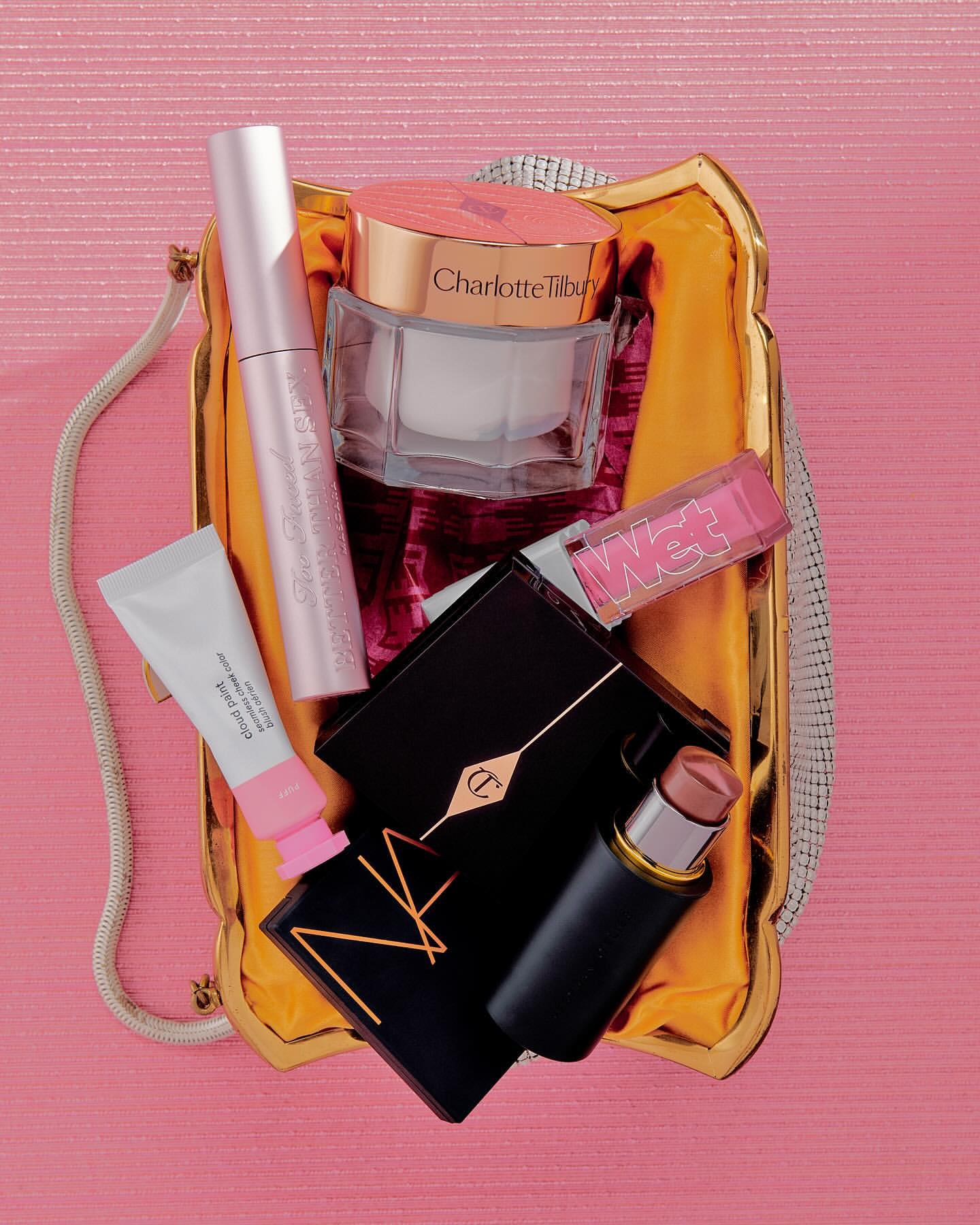 Close-up of makeup products in a gold handbag against a pink backdrop: Charlotte Tilbury, Nars, Too Faced, Glossier, and Wet n Wild.