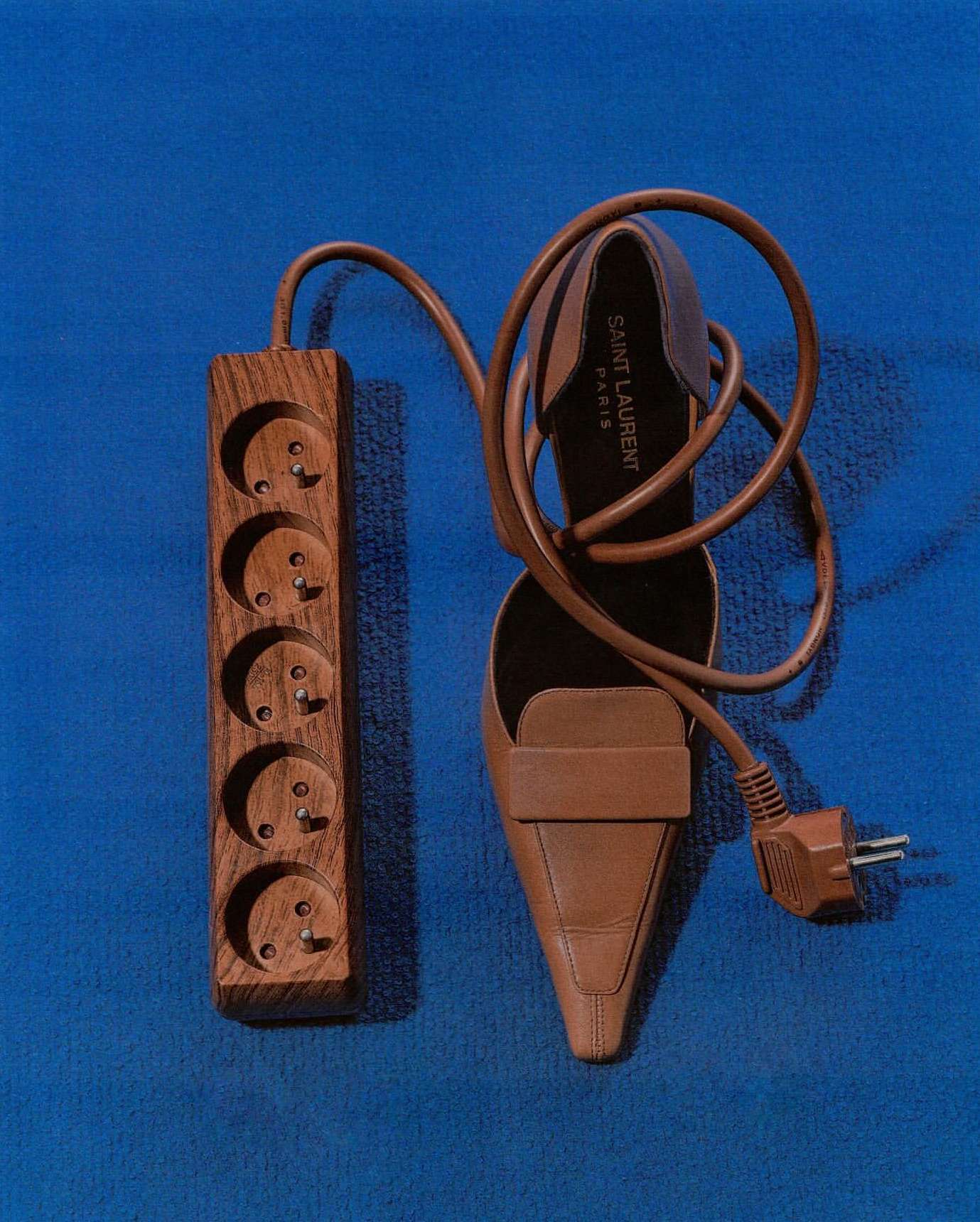 Still life shot of a brown leather stiletto heel, an electrical cord, and a wooden power strip against a blue background.