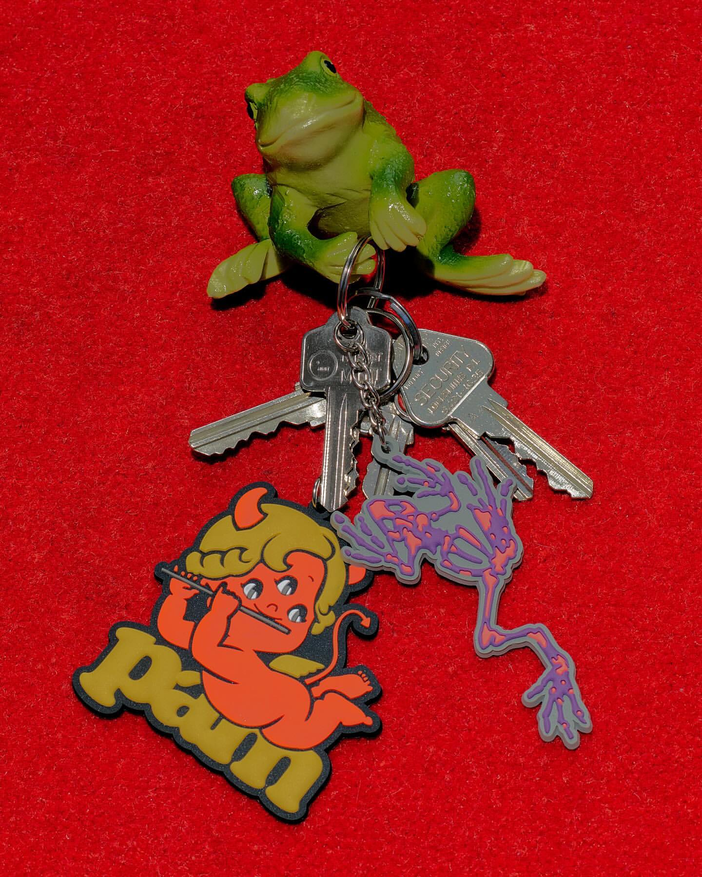 A green frog figurine sits atop a keyring holding silver keys and colorful cartoon charm attached against a textured red surface.