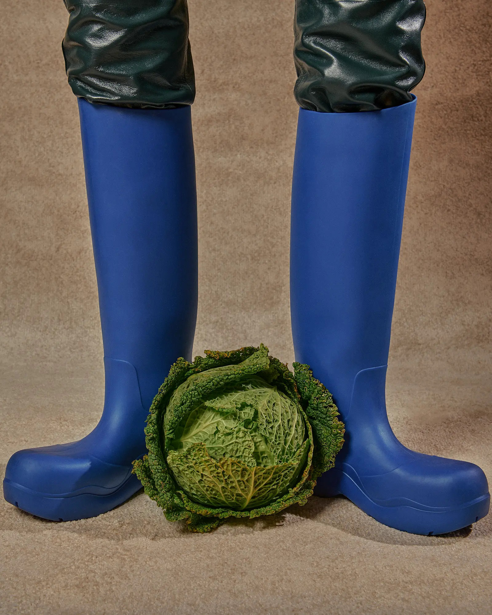 Eye-level shot of blue rubber boots worn with green waterproof material, paired with a green savoy cabbage on a neutral background.