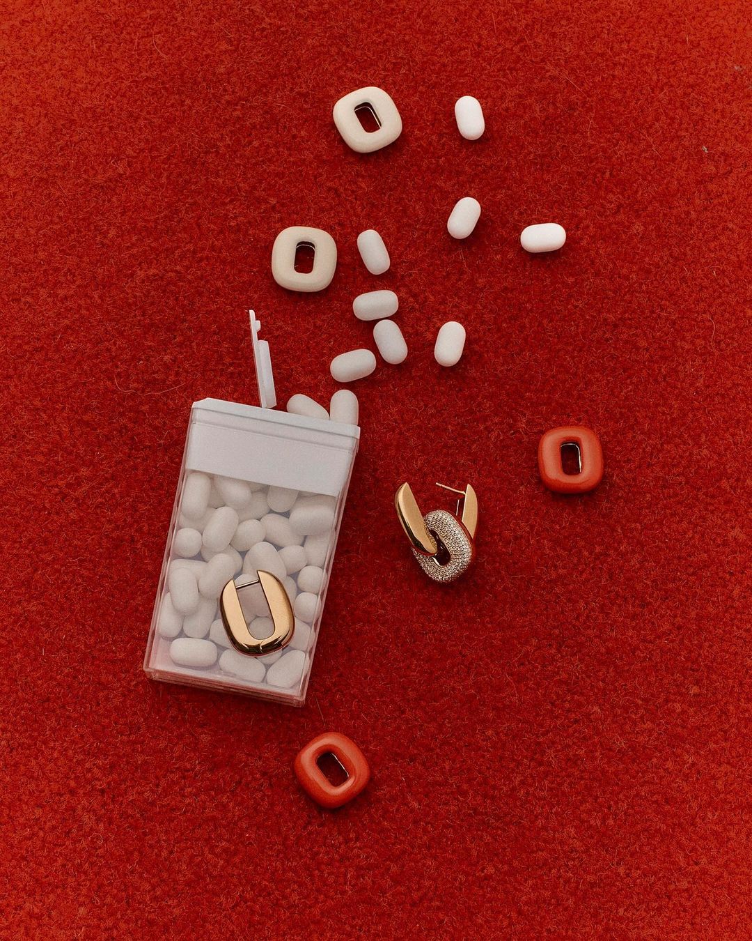 Still life flat lay: earrings, mints, and colorful shapes are arranged on a red, textured surface, creating a visually appealing composition.