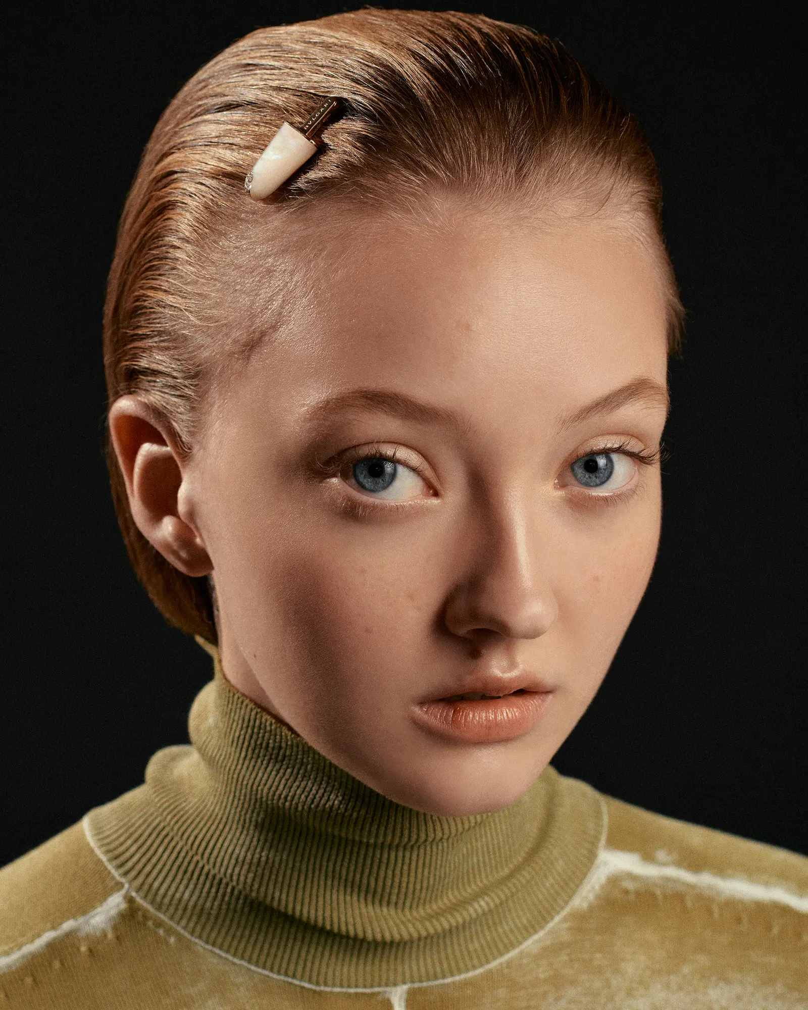 Close-up portrait of a fair-skinned adult woman with slicked back reddish-blonde hair, light eyes, and a neutral expression against a dark backdrop.