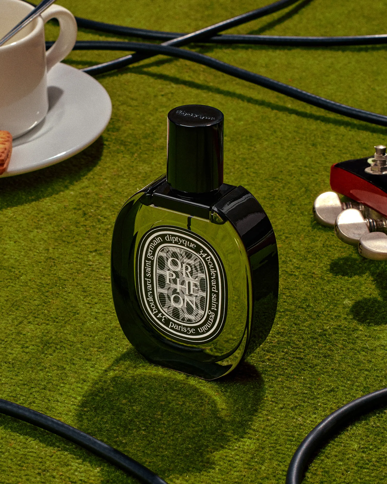 Overhead studio shot of a Diptyque Orpheon perfume bottle on a green surface with a teacup, guitar part, and cables.