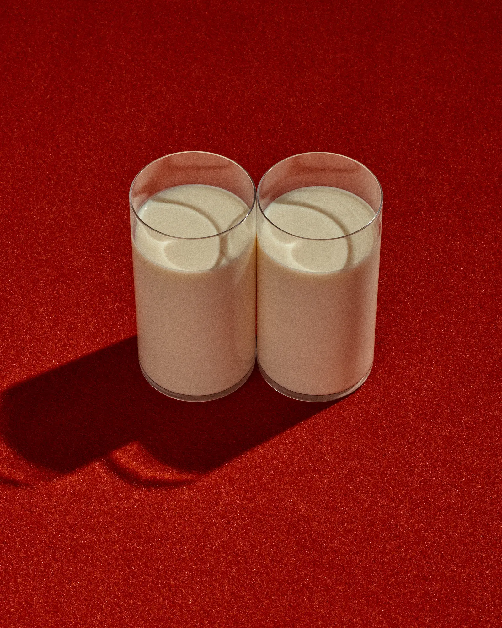 Two glasses filled with creamy white milk stand side-by-side on a textured red background, casting distinct shadows in a brightly lit scene.