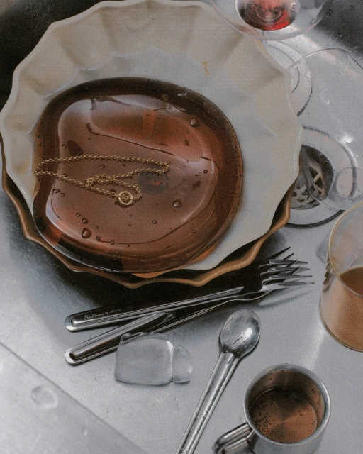 Still life of a brown plate with a gold necklace, forks, spoons, and a metal mug in a stainless steel sink with running water.