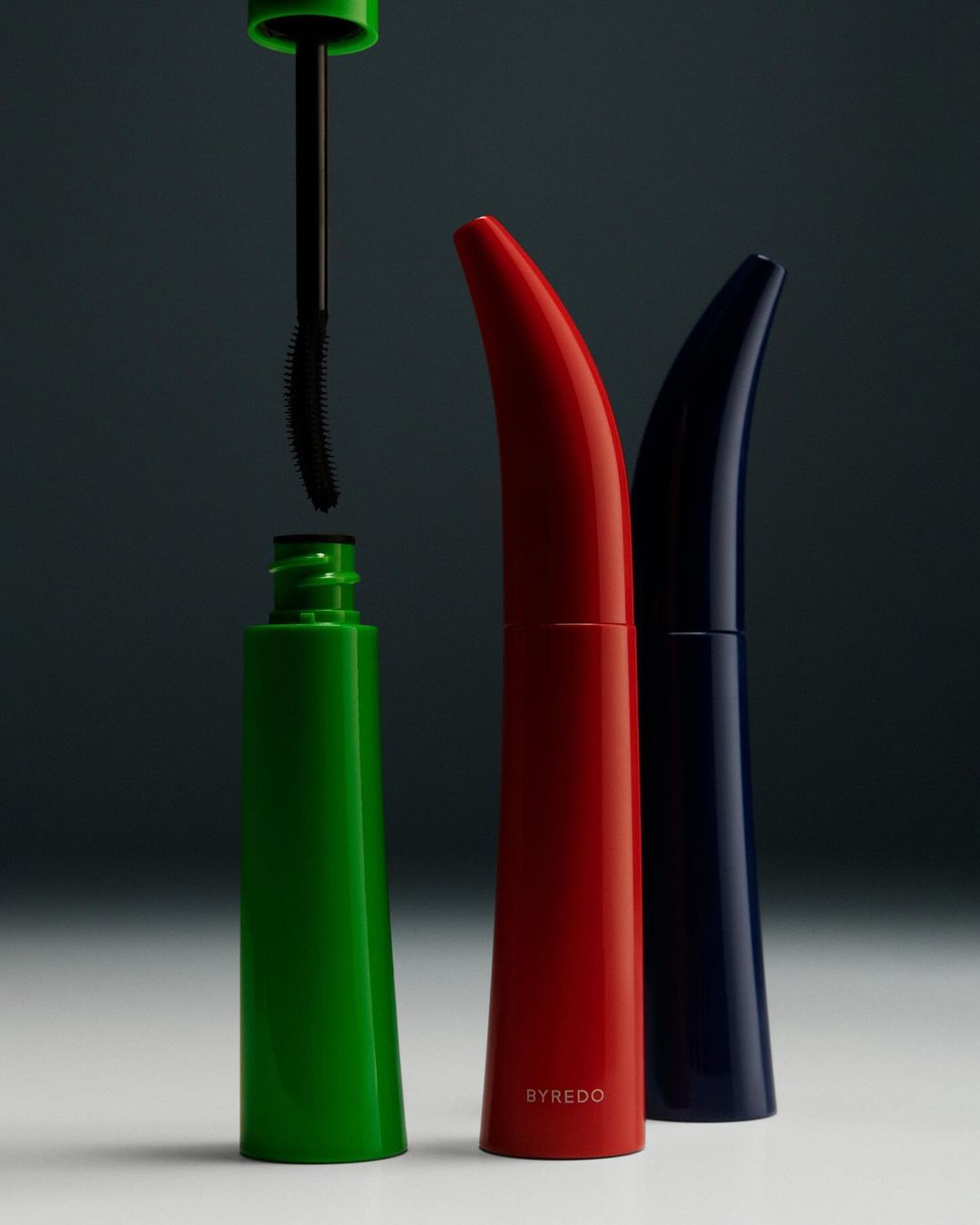 Still life shot of three colorful Byredo mascara containers, one green with applicator, plus red and blue, on a graduated gray background.