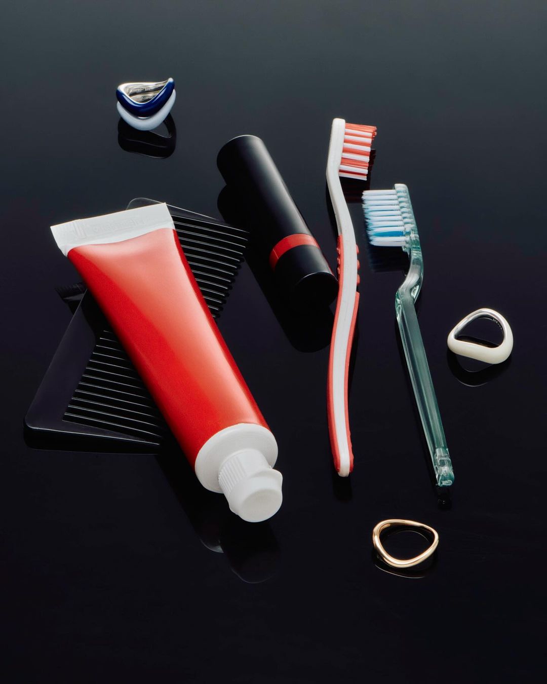 Still life studio shot of a red toothpaste tube, black lipstick, toothbrushes, dental flossers, and a black comb on a black background.