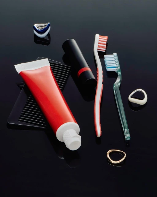 Still life studio shot of a red toothpaste tube, black lipstick, toothbrushes, dental flossers, and a black comb on a black background.