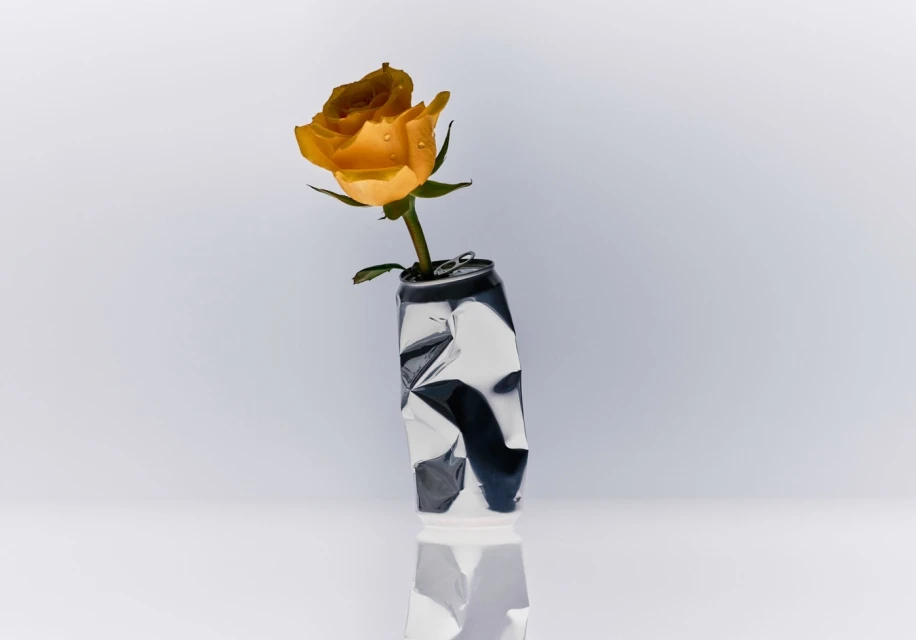 Still life of a single yellow rose with water droplets in a silver crushed aluminum can, placed on a white surface against a white background.