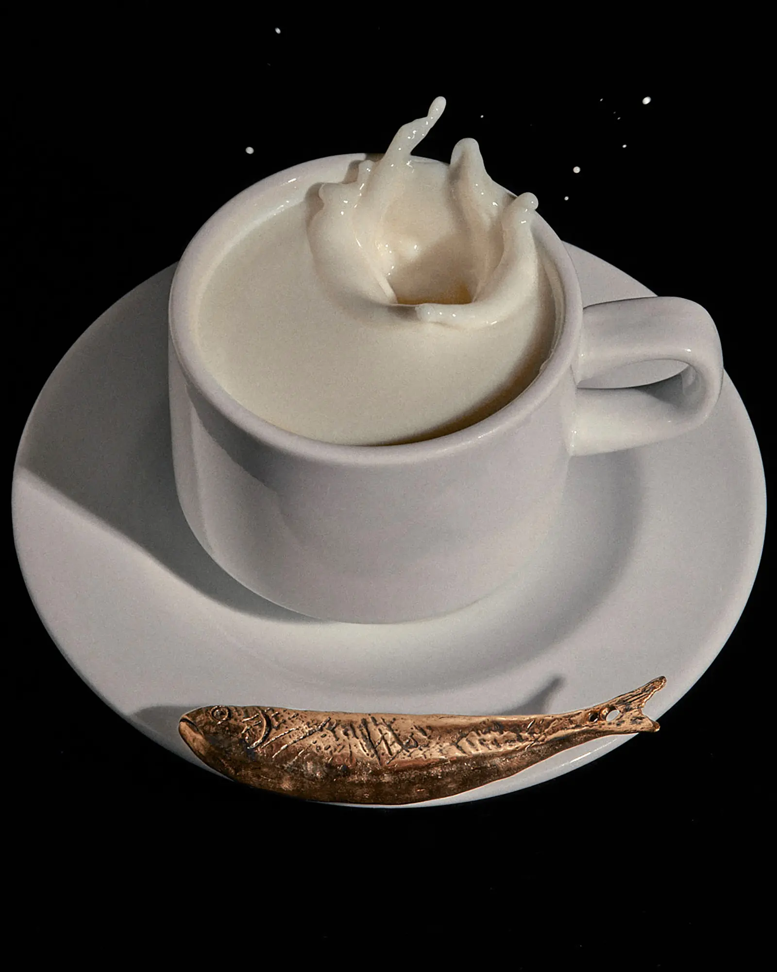 Close-up still life showcasing milk splashing in a white cup, set on a saucer with a bronze-colored fish figurine against a black background.