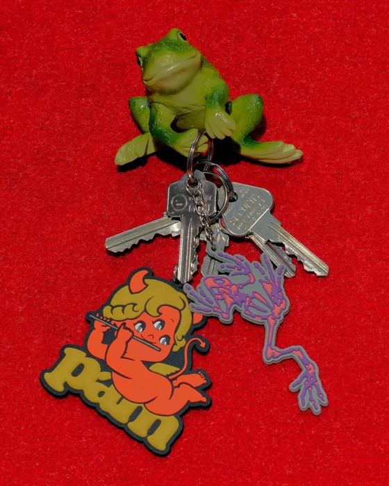 A keyring featuring a green plastic frog holding metal keys, a red demon charm, and a purple skeletal hand charm on a red felt background.