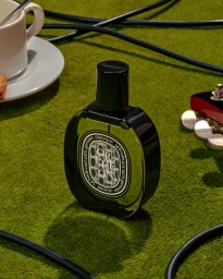 Black perfume bottle on green textured grass with white coffee cup and guitar tuning pegs nearby