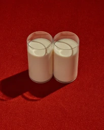Two tall glasses filled with white milk placed side by side on a textured red surface, casting long shadows.
