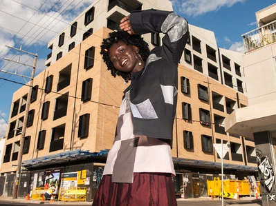 A woman in a patterned jacket smiles with a hand raised, posing in an urban setting with a modern building and blue sky backdrop.
