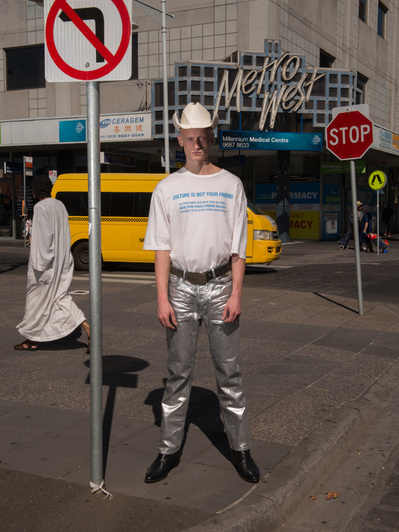A person in shiny silver pants and a cowboy hat poses on a city street next to a no left turn sign and stop sign, with a bus in the background.