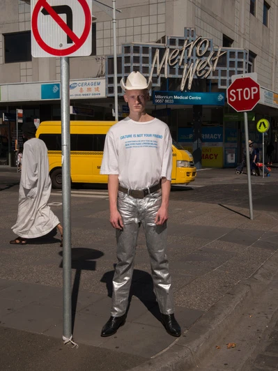 A person in shiny silver pants and a cowboy hat poses on a city street next to a no left turn sign and stop sign, with a bus in the background.