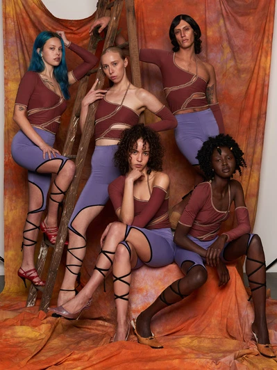 Six diverse adult fashion models pose in matching brown crop tops and periwinkle bottoms against an orange-tinted canvas backdrop indoors.