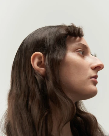 Profile portrait of a woman with long wavy brown hair and styled bangs against a plain light background