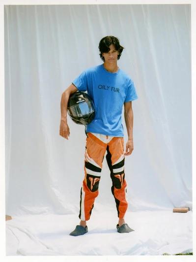 Young adult poses in a blue tee and motocross pants, holding a motorcycle helmet against a white backdrop, fashion editorial.