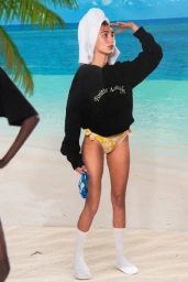 Model with white towel on head wearing black sweatshirt and yellow bikini bottoms standing against painted beach backdrop