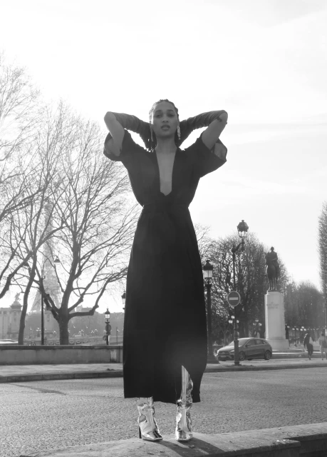 Woman in long black dress and metallic boots poses outdoors near trees and statues, with the Eiffel Tower visible in the background.